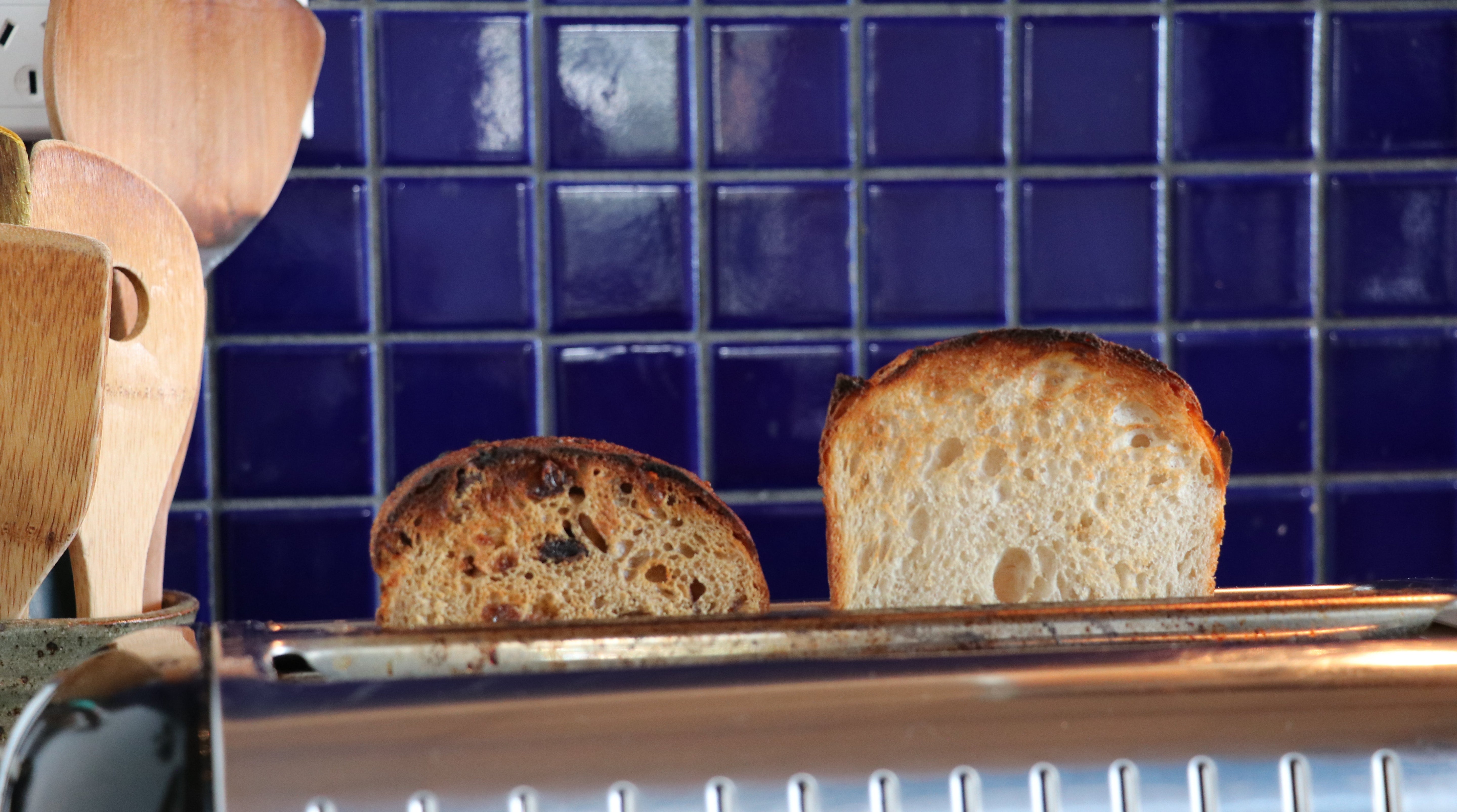 Two slices of toasted bread emerging from a stainless steel toaster in a kitchen setting, with a dark blue tiled backsplash behind it.