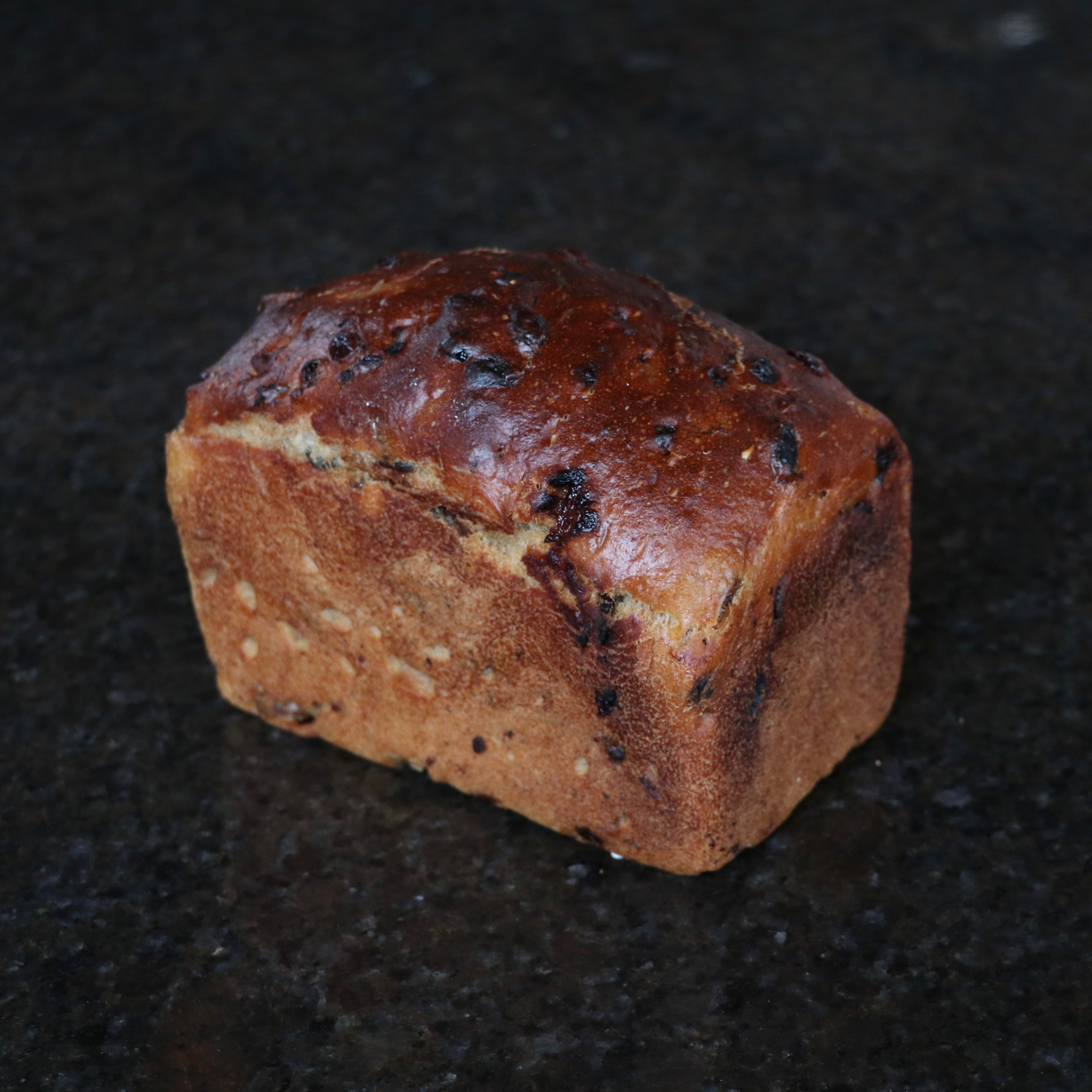A single loaf of rustic bread with a glossy dark brown top and golden-brown sides, photographed on a dark speckled countertop.