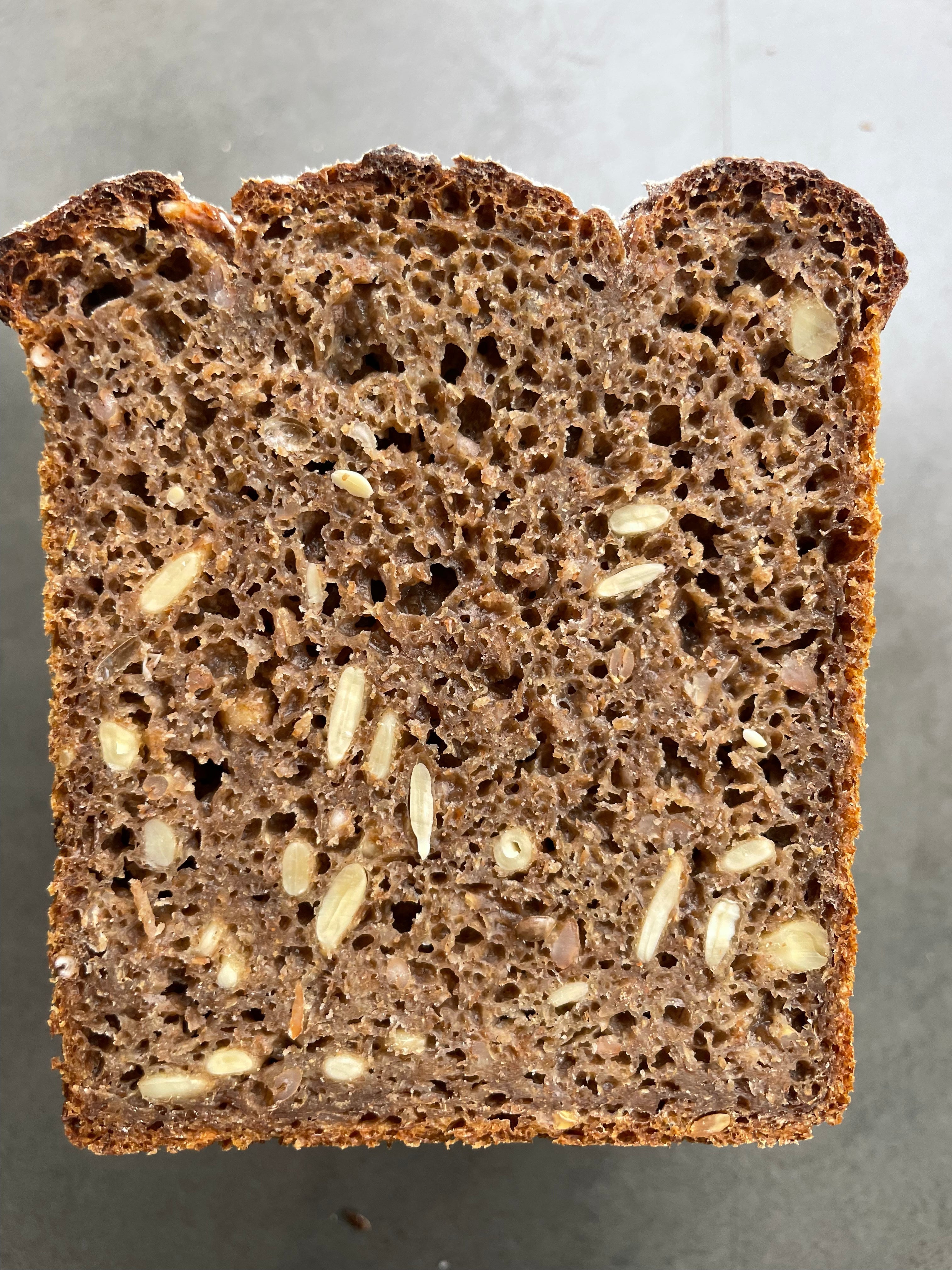 A thick slice of dark brown rye bread with visible sunflower kernels, shown from a top-down view on a neutral gray surface.